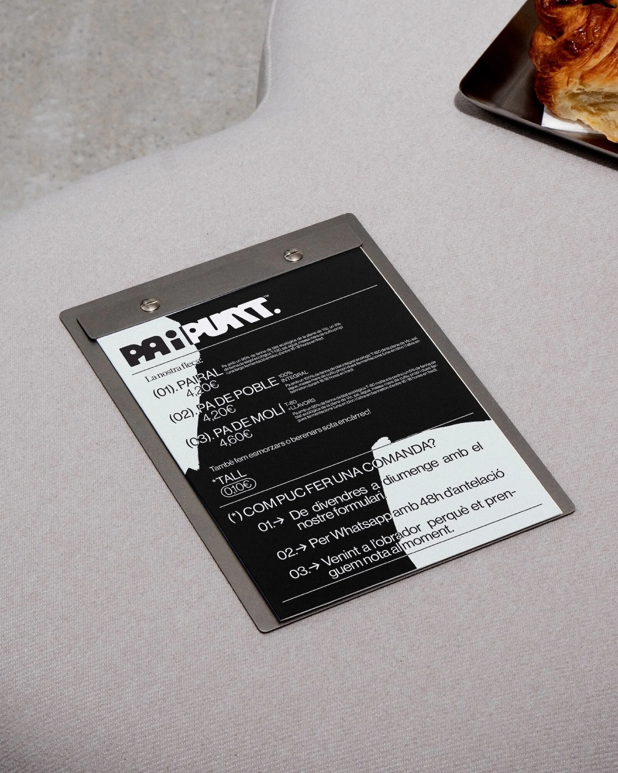 Close-up of a cafe menu on a metal clipboard showing menu items and prices, placed on a light grey surface.