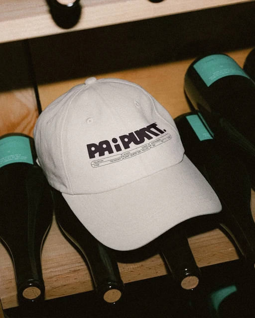 Close-up of a light grey baseball cap with black lettering on top of black wine bottles in a wooden wine rack indoors.