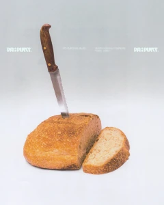 Bread knife stuck vertically into a loaf of rustic bread with one sliced piece resting beside it against a plain white background.