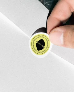 Close-up of fingers peeling a circular yellow branded sticker with black typography off a white envelope.