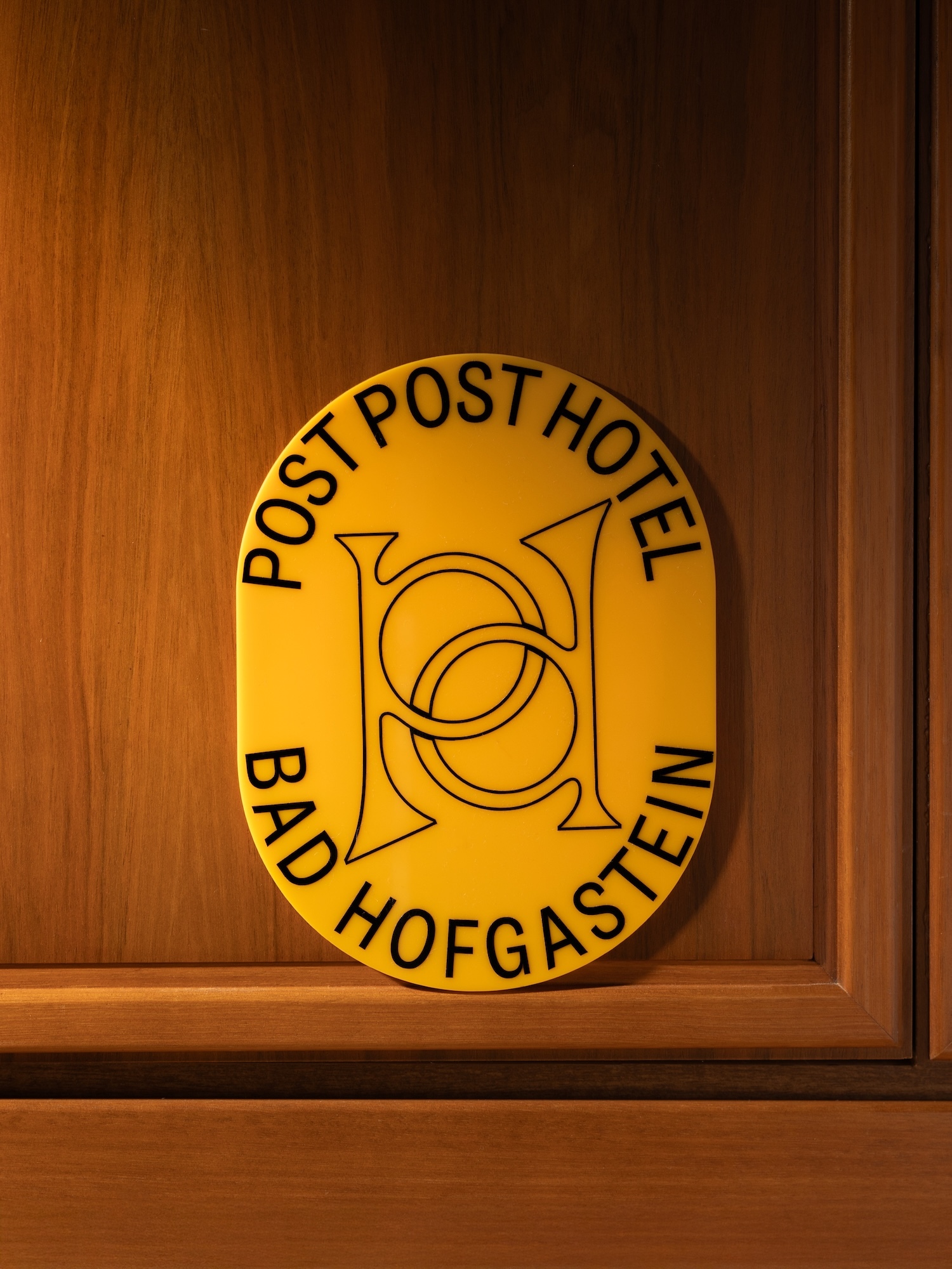 Close-up of a yellow oval hotel sign reading 'Post Post Hotel Bad Hofgastein' with a black line art logo, mounted on a wooden wall.