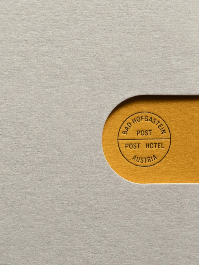 Textured off-white paper featuring a die-cut window revealing vibrant yellow paper with a circular stamped Post Hotel crest.