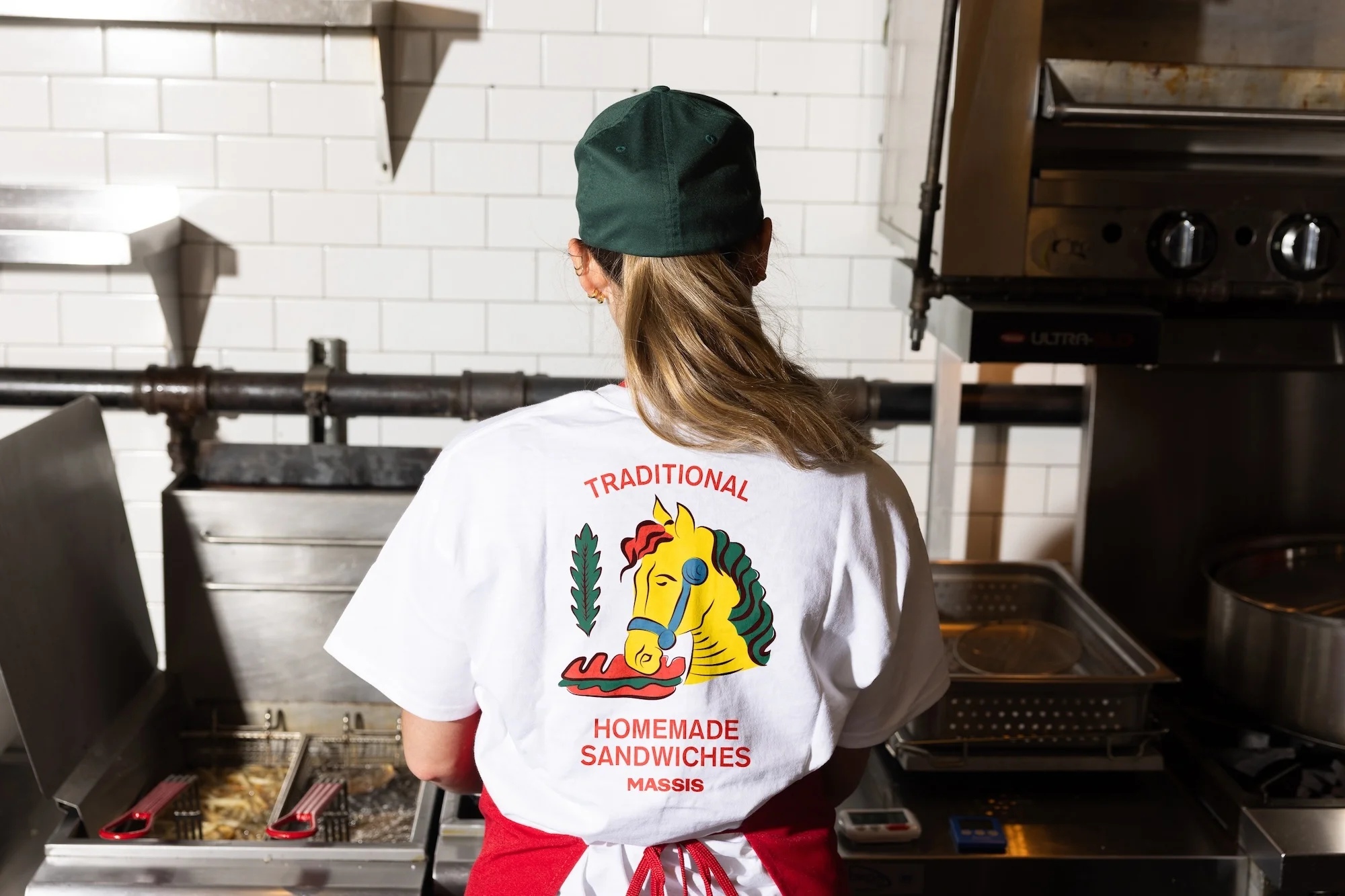 Back view of a woman in a kitchen wearing a t-shirt with a horse and sandwich graphic, ready to cook near restaurant equipment.
