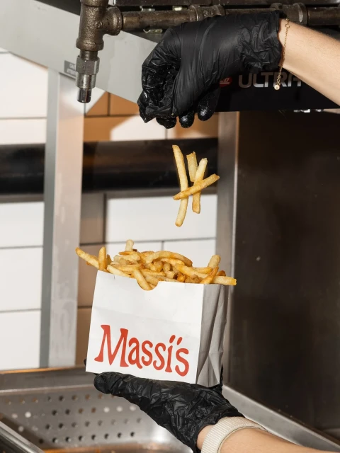French fries being poured from a paper bag held by hands in black gloves with an industrial background, indoors, and food presentation.