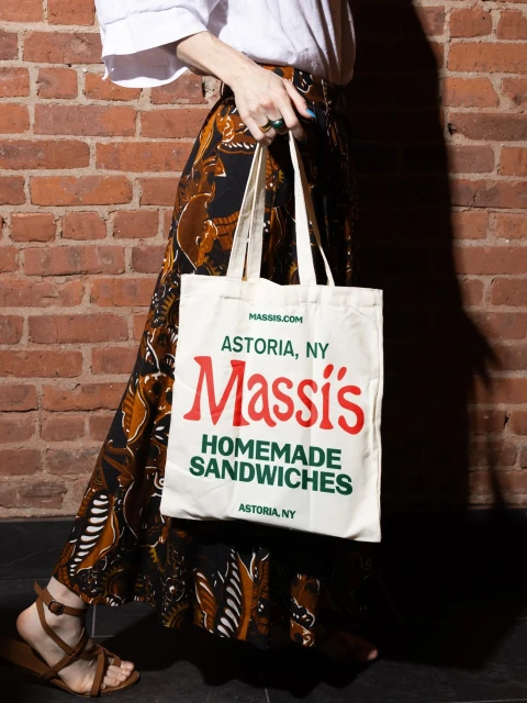 Adult female walks beside brick wall carrying white tote bag with 'Massis Homemade Sandwiches Astoria, NY' printed in red and green.