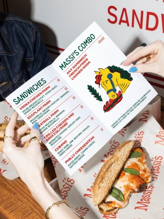 Overhead shot of a Massi's Combo menu in hands, next to a sandwich on a branded paper, capturing a culinary scene indoors.