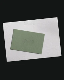 A sage green business card with a debossed M logo resting on top of a larger white textured paper sheet against a dark background