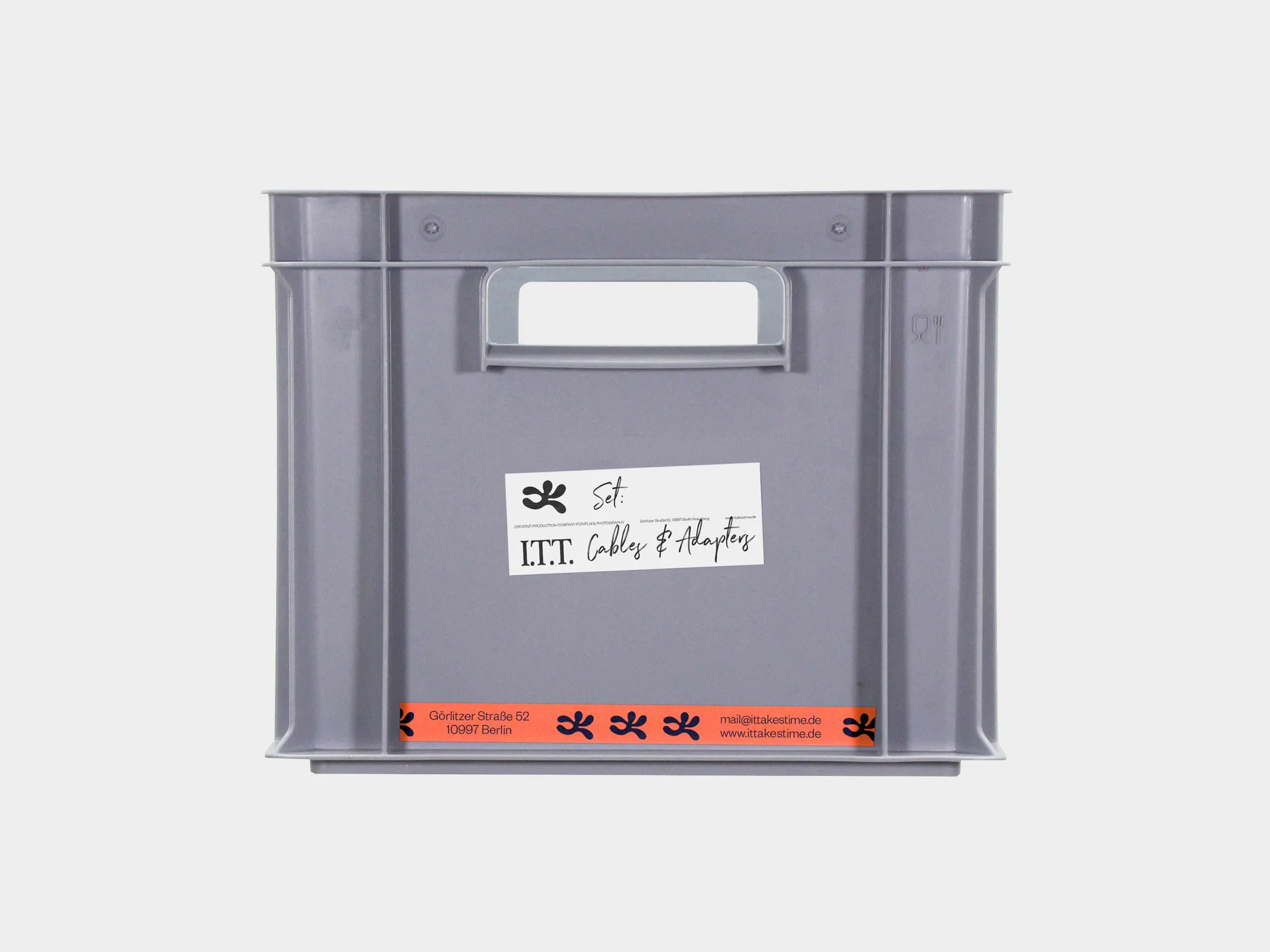 Grey plastic crate with handles and a label on a white background, featuring address and contact details, and text reading 'Set: Cables & Adapters'.