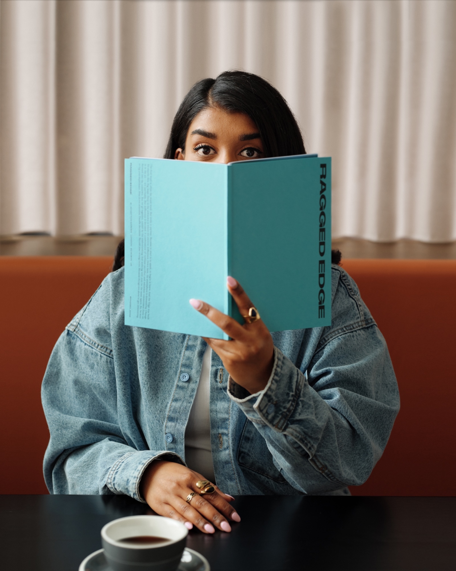 Young woman sits at table, reading a book titled 'Ragged Edge.' She wears a jean jacket with rings and a cup of coffee is nearby.