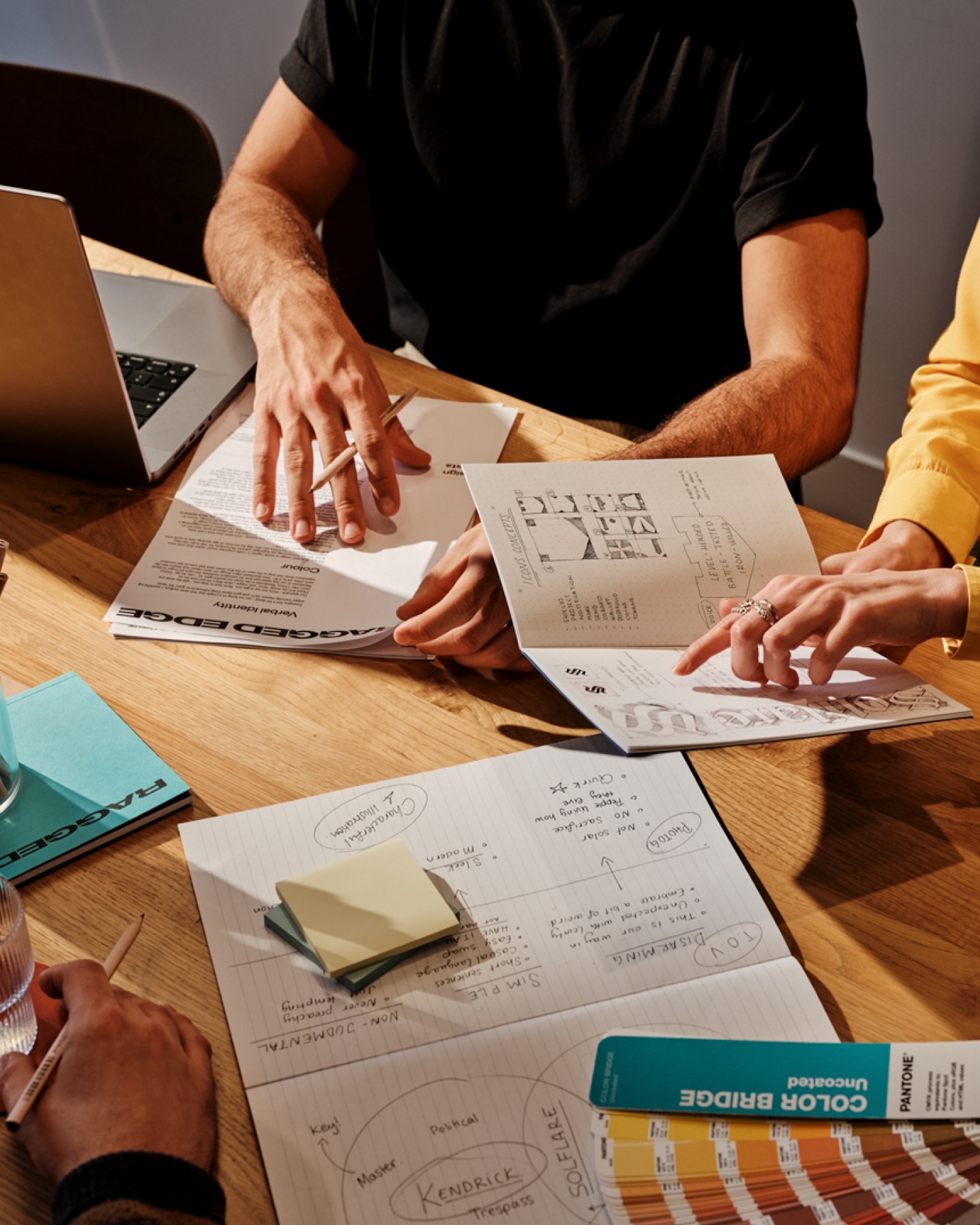 Overhead shot of three adults collaborating on documents and color palettes on a light wood table with a laptop.