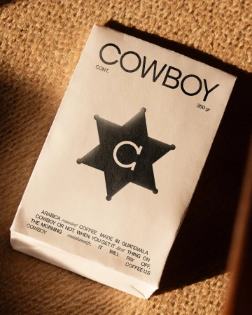 Close-up product shot of a white coffee bag with 'COWBOY' printed on it in black ink, set against a tan woven burlap surface.