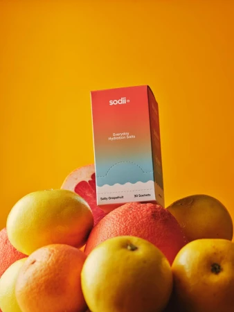 Box of Sodi Everyday Hydration Salts sitting atop pile of yellow and red grapefruits against bright yellow background in a studio shot.