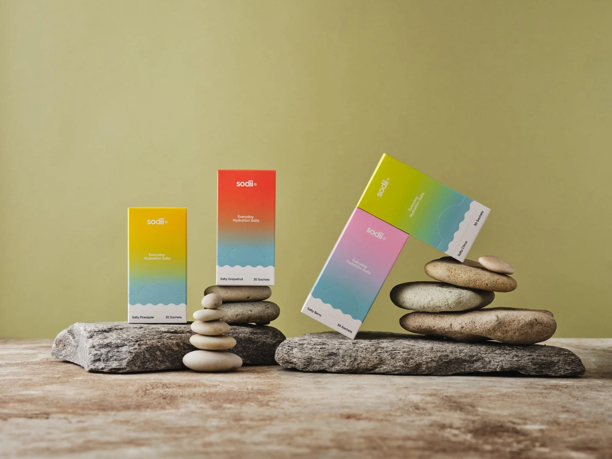 Close-up shot of colorful sodi+ hydration salts boxes on stone platforms, arranged with small rocks, set against a pale green backdrop.