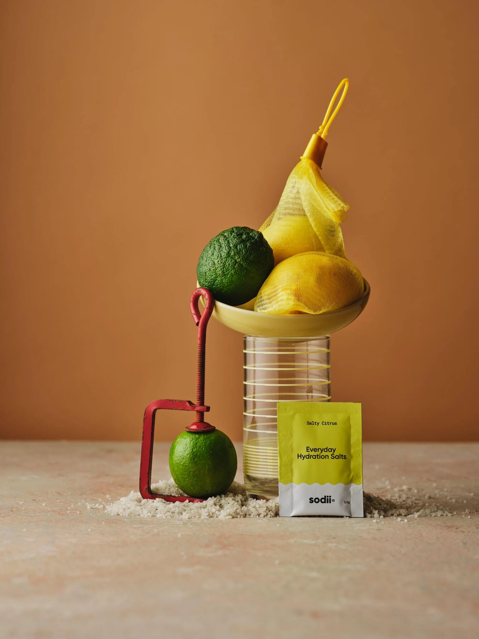Still life photograph of lemons and limes in a yellow mesh bag, scale, and hydration salts against a tan and pink background.