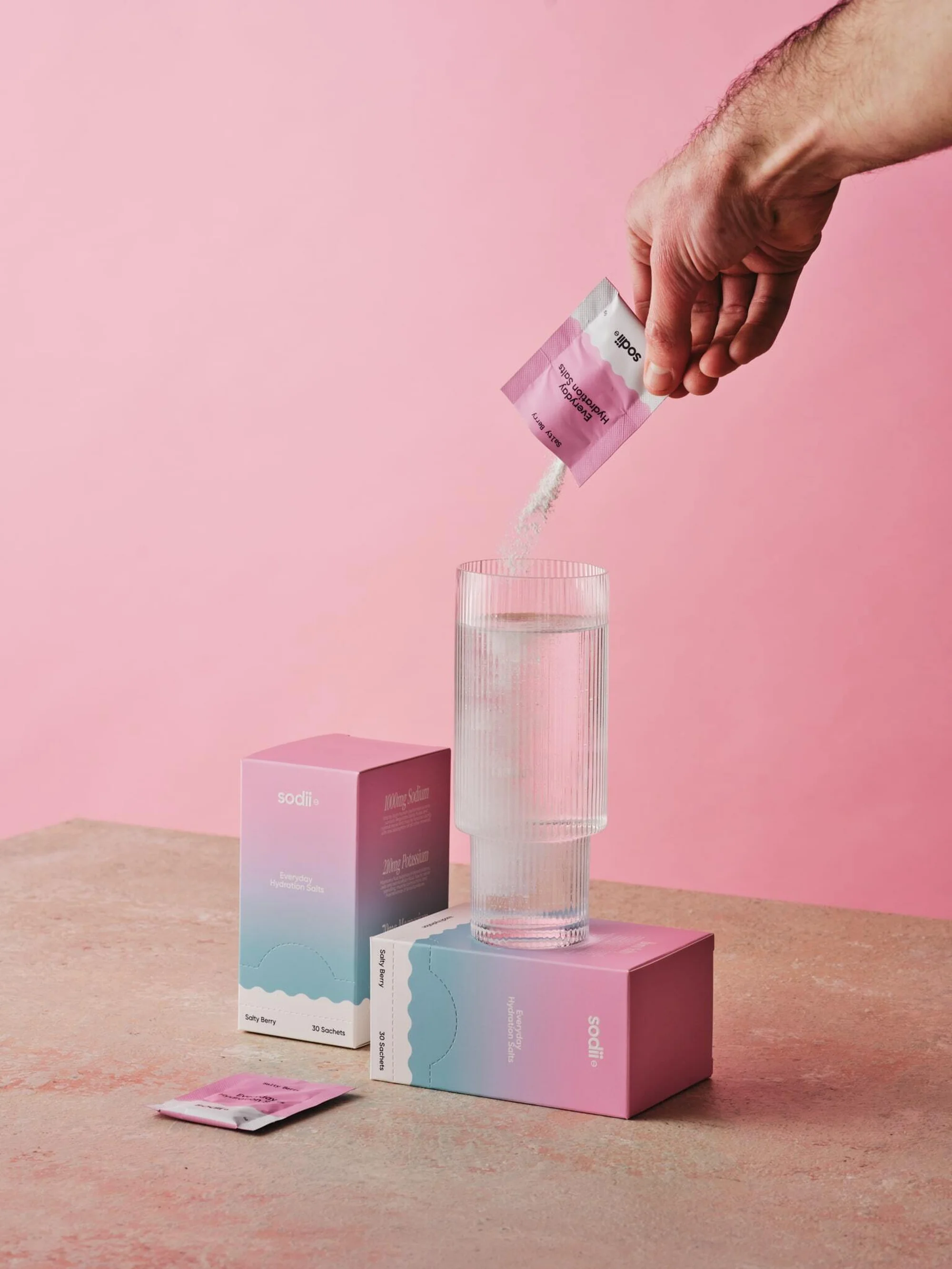 Arm pours white powder from sachet into tall glass of water, resting on product boxes, against pink backdrop in studio setting.