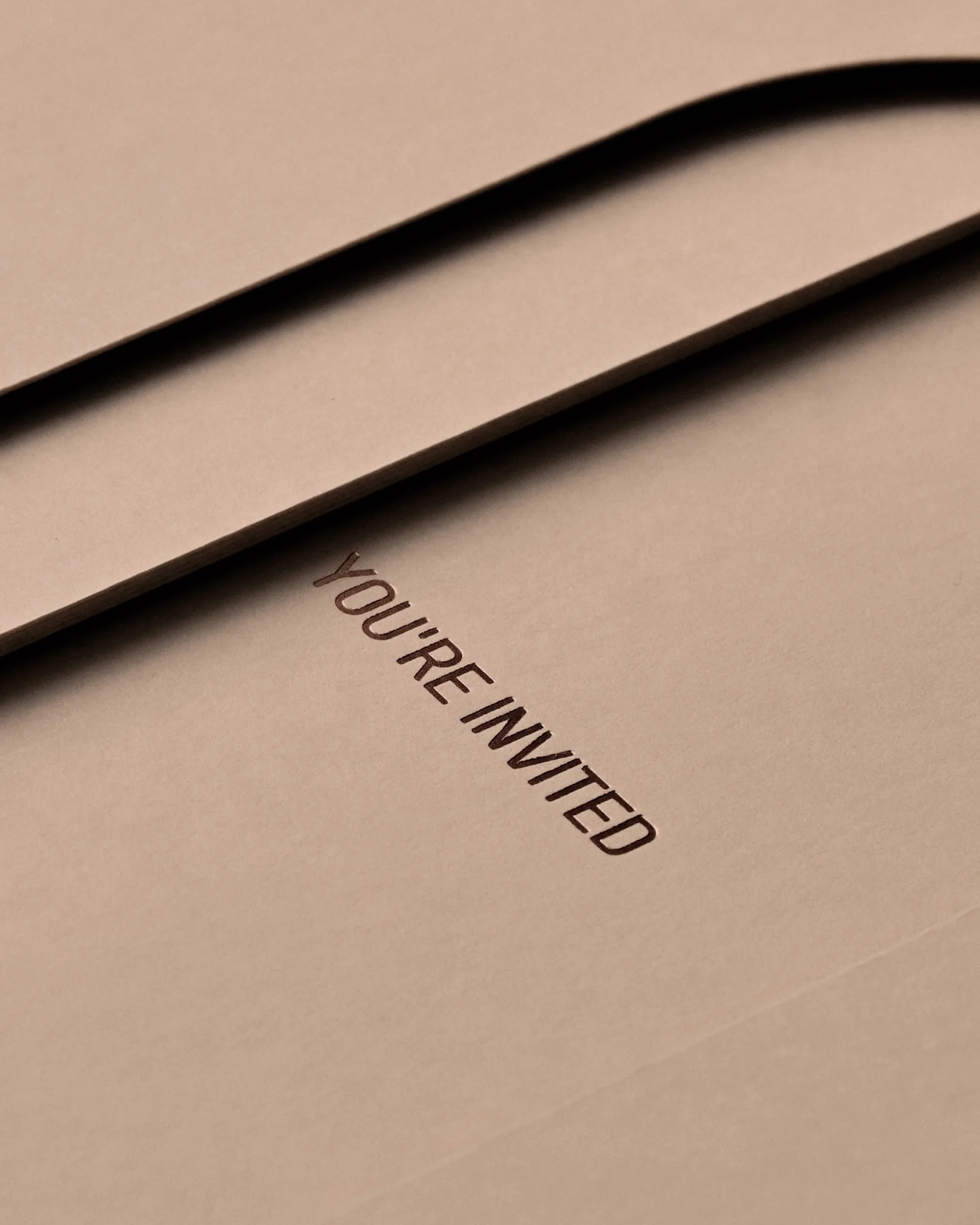 Close-up of an invitation with the words 'You're Invited' printed on a blush-colored card with a dark, rounded band.
