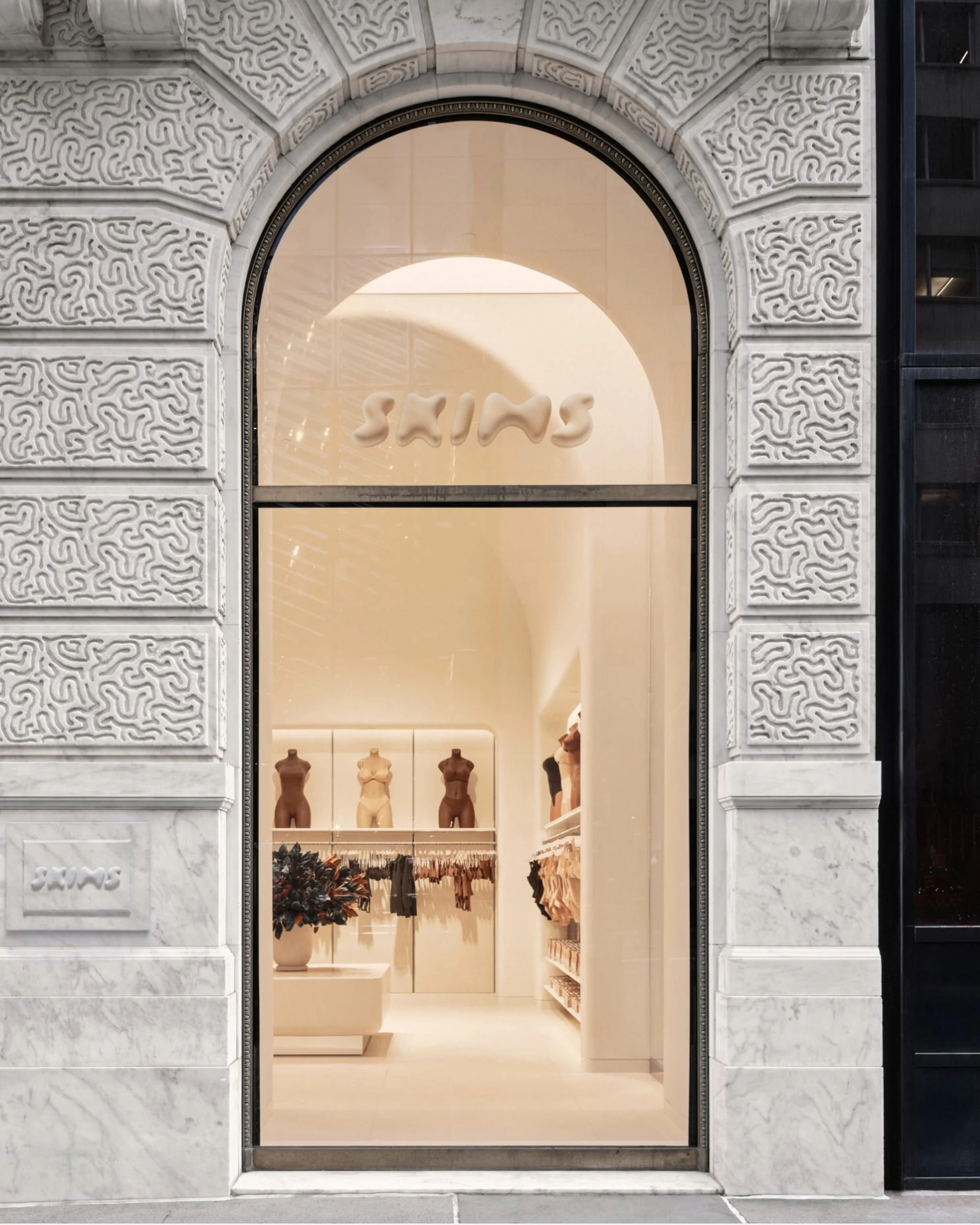 The SKIMS retail store, showcasing clothing on mannequins and shelves visible through the arched glass exterior storefront in an urban setting.