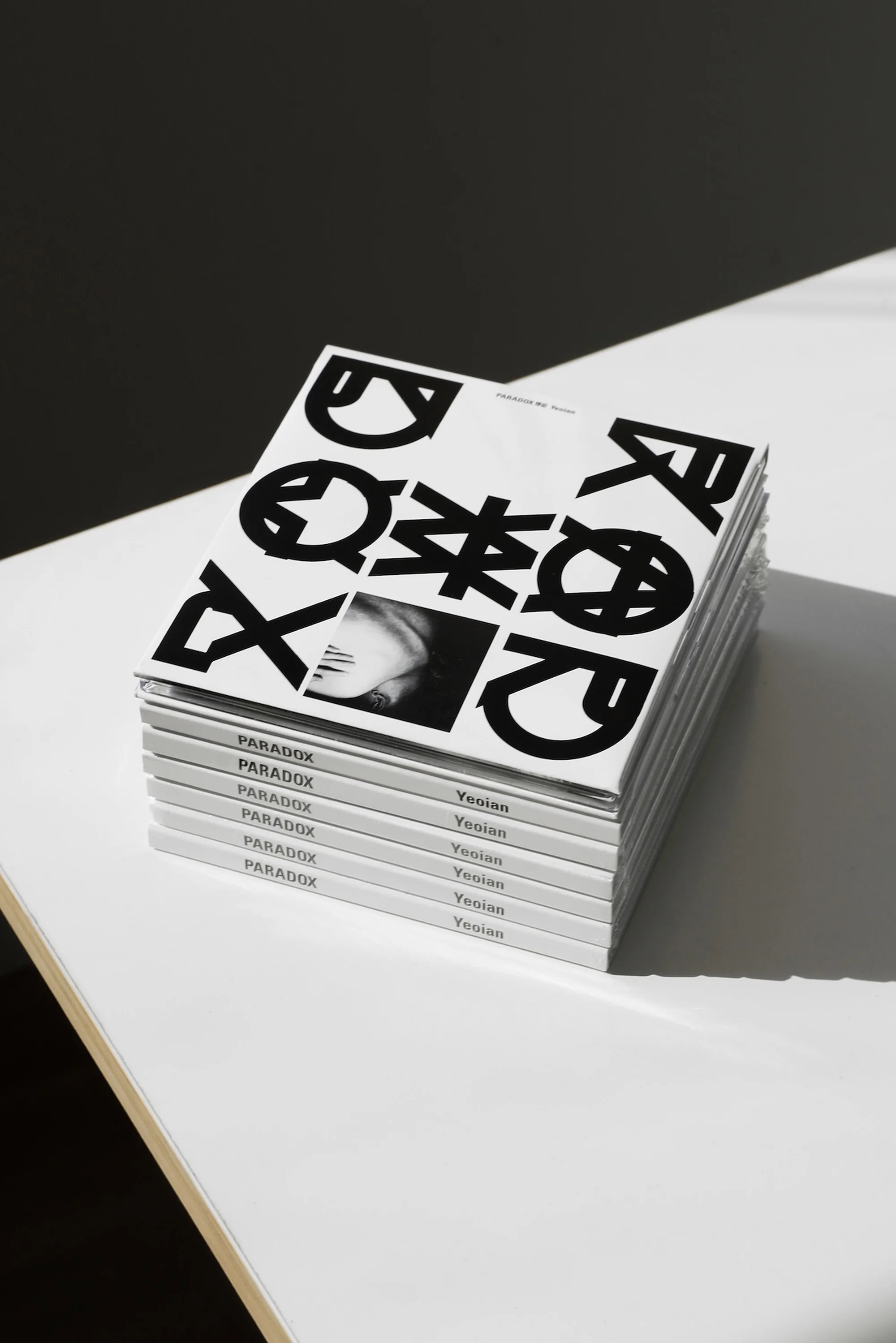 Stack of Paradox magazines on a white table with geometric black and white cover design, and bright window lighting.