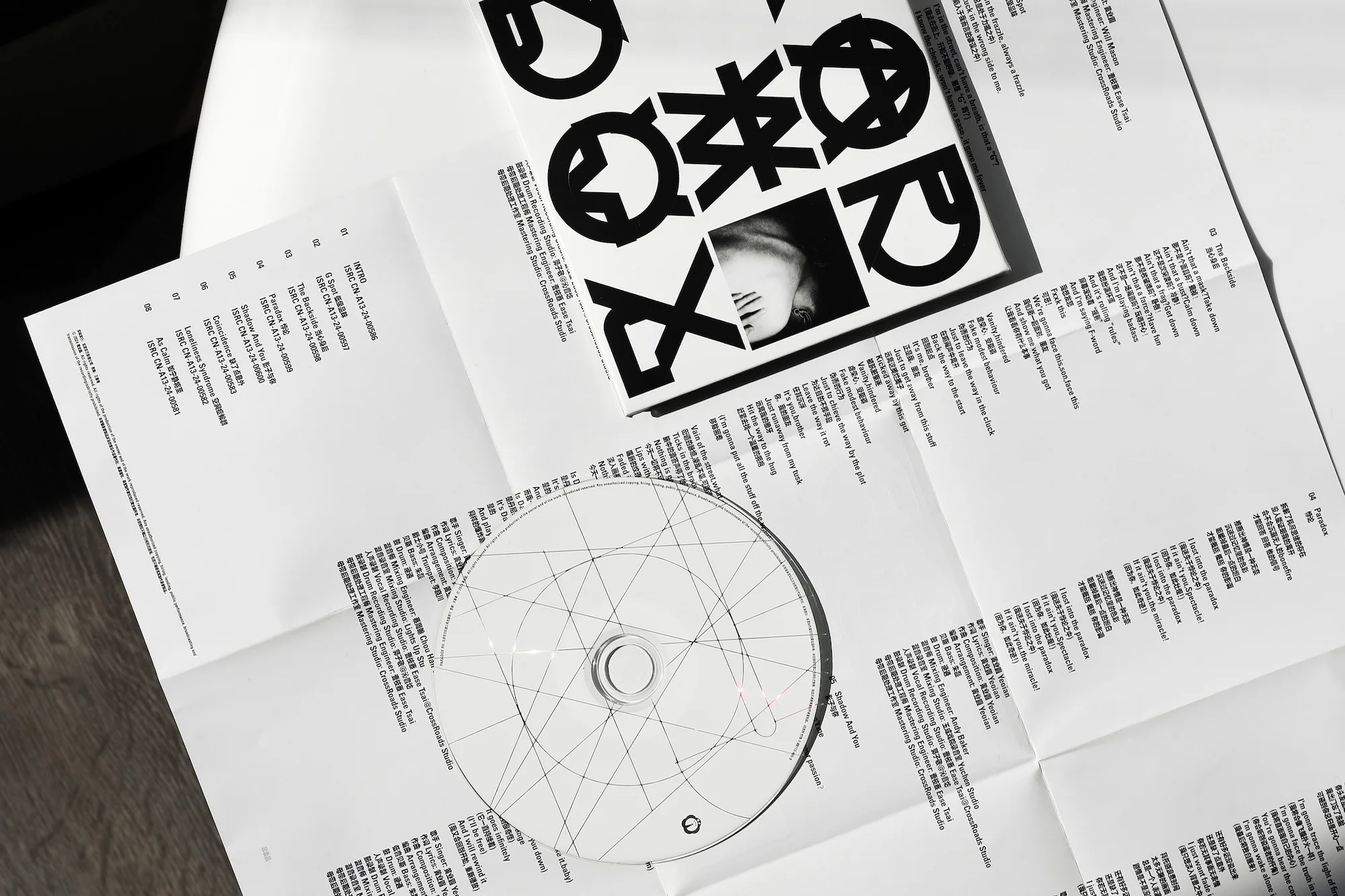 Overhead shot of a CD, a booklet with a woman's face, and pages of track listings on a white surface. Black and white aesthetic.