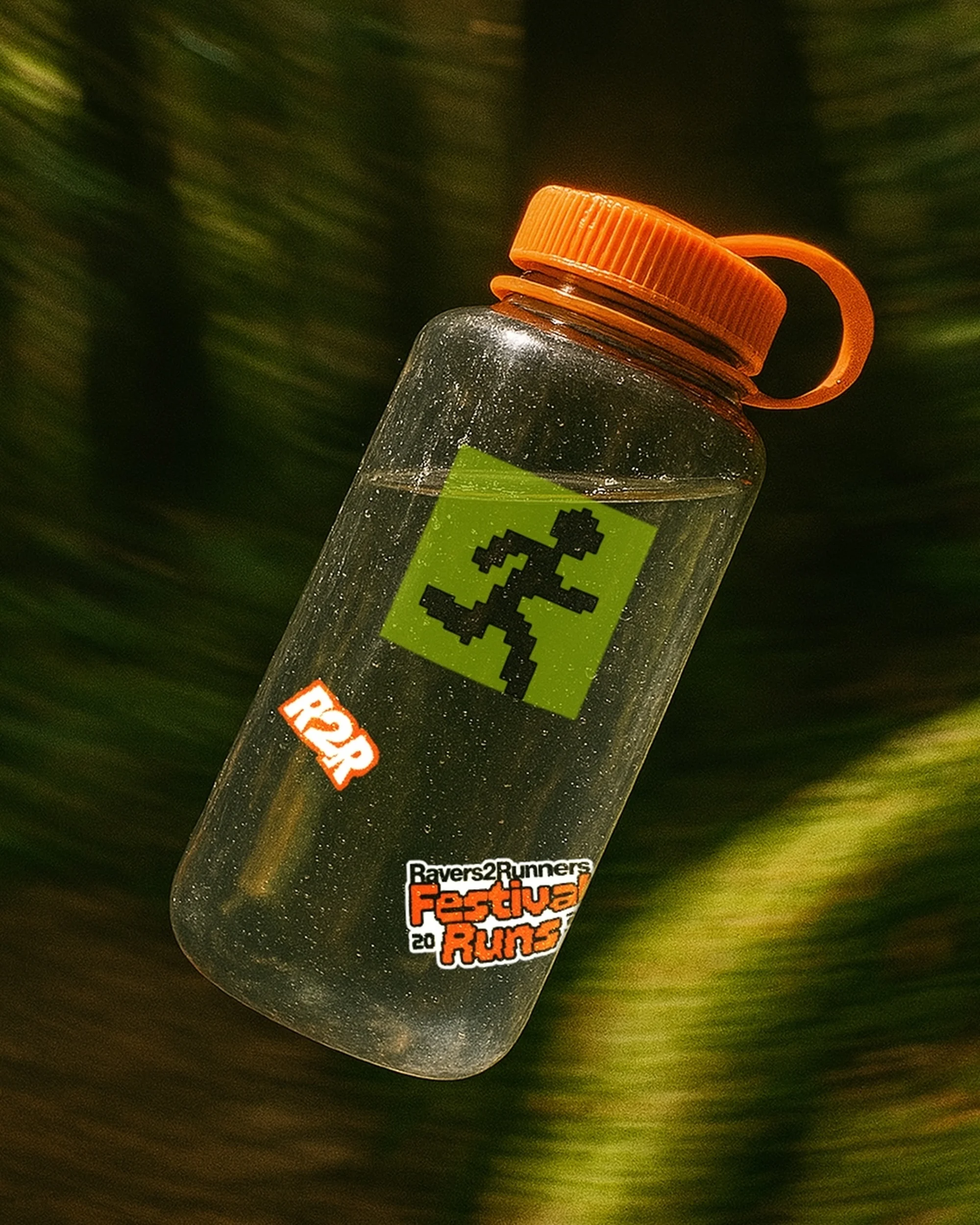 Clear plastic water bottle with orange lid, covered in stickers, floats in front of a blurry green leafy background in soft light.