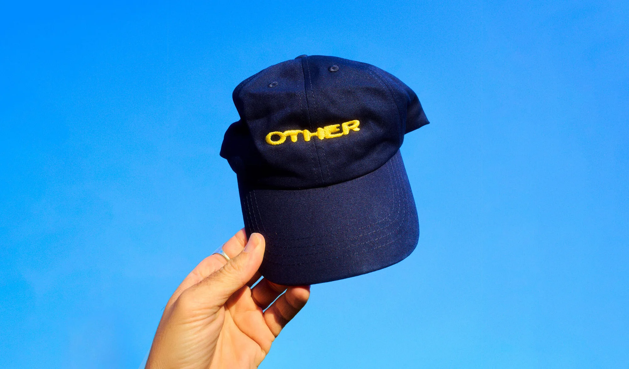 Hand holding a dark blue baseball cap with 'OTHER' embroidered in yellow against a clear, vibrant blue sky backdrop.