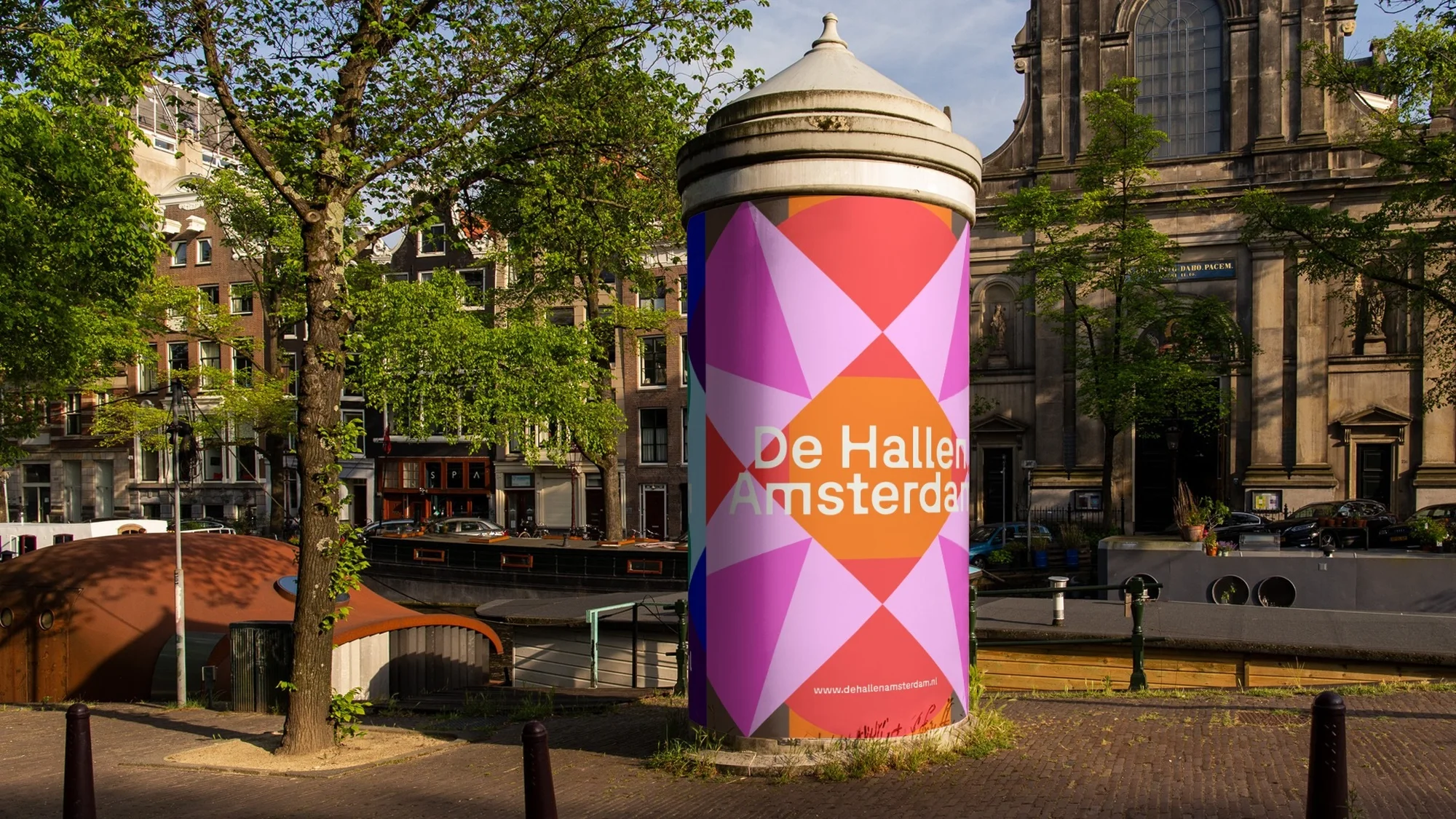 Colourful advertising column for De Hallen Amsterdam featuring a geometric design, set against the backdrop of Amsterdam architecture.