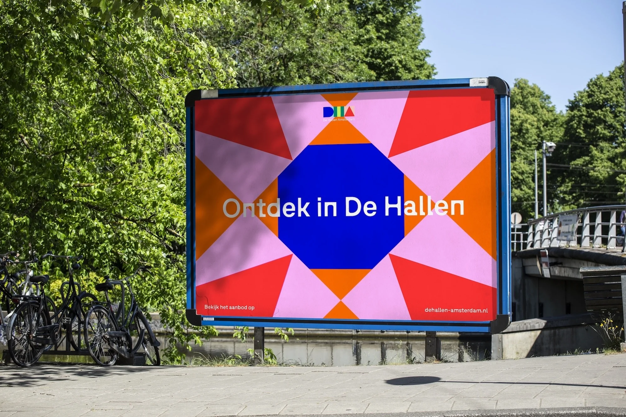 Outdoor billboard featuring an abstract geometric design with text "Ontdek in De Hallen", surrounded by trees and parked bicycles.