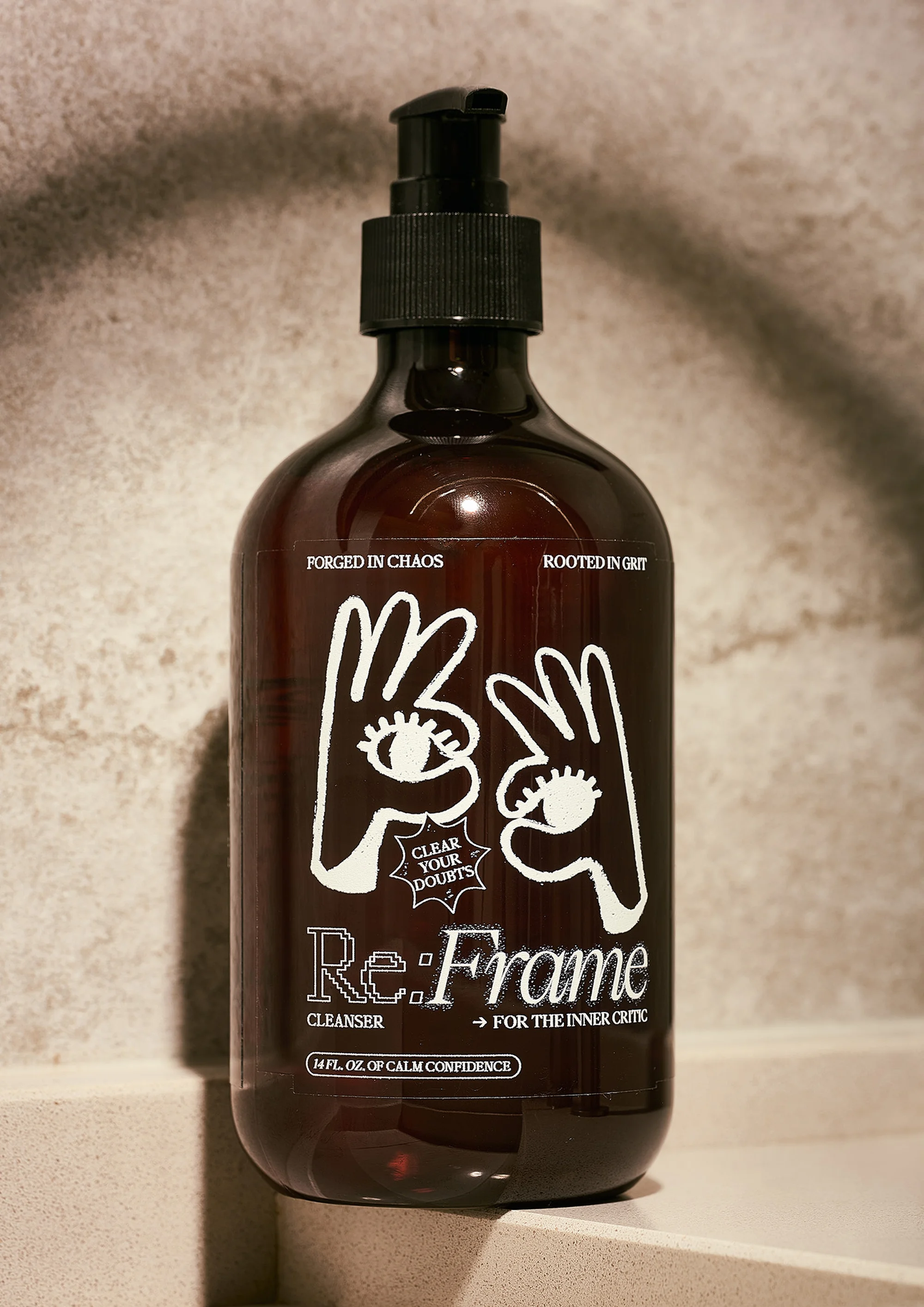 Close-up of a brown glass bottle of Re:Frame cleanser with black pump dispenser. Bottle features white artistic text and hand graphics.