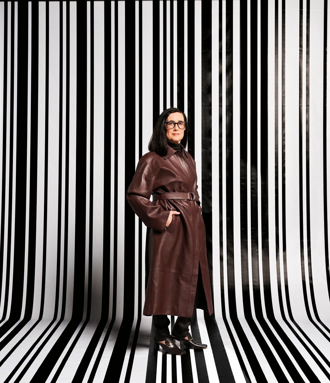 A stylish woman in a brown leather coat stands in front of a black and white striped background in a full portrait shot.