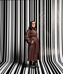 Gina Cass-Gottlieb wearing a brown leather coat standing against a black and white striped background