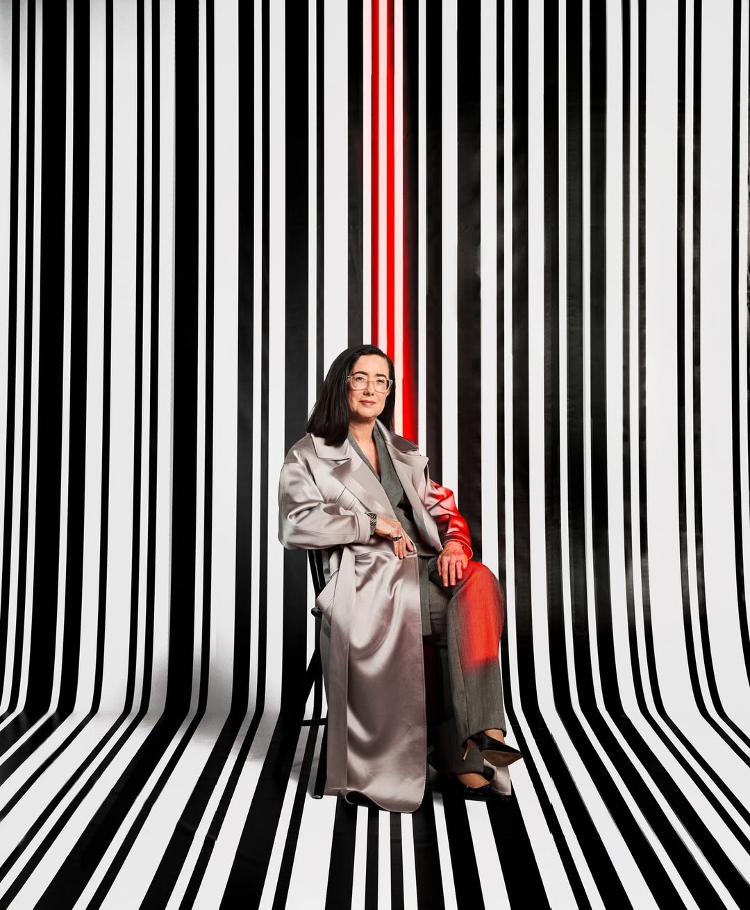 Elegant woman in a silver coat sitting in a studio with a black and white striped background and a red stripe, creating a striking portrait.