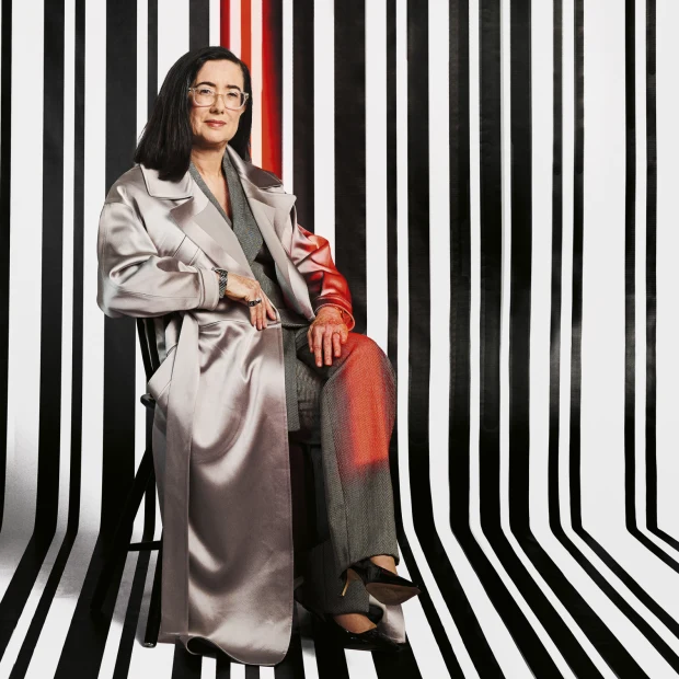 Elegant woman with glasses poses in a shiny silver coat and gray suit, seated in front of a black and white striped backdrop.
