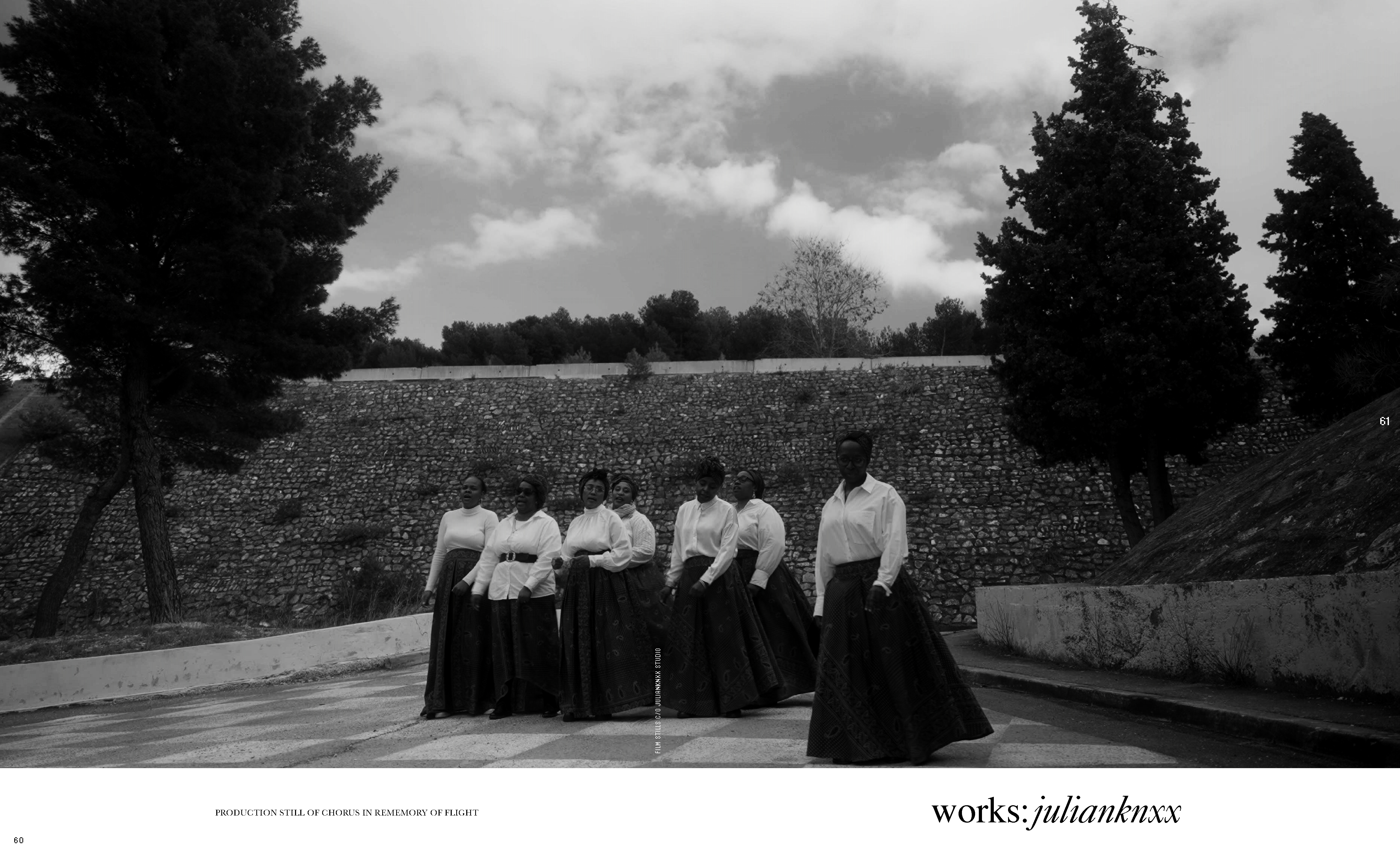 Seven women of African descent stand on a stone path in formal attire—white tops and dark skirts—against a stone wall and trees under a cloudy sky.