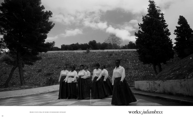 Seven women of African descent stand on a stone path in formal attire—white tops and dark skirts—against a stone wall and trees under a cloudy sky.