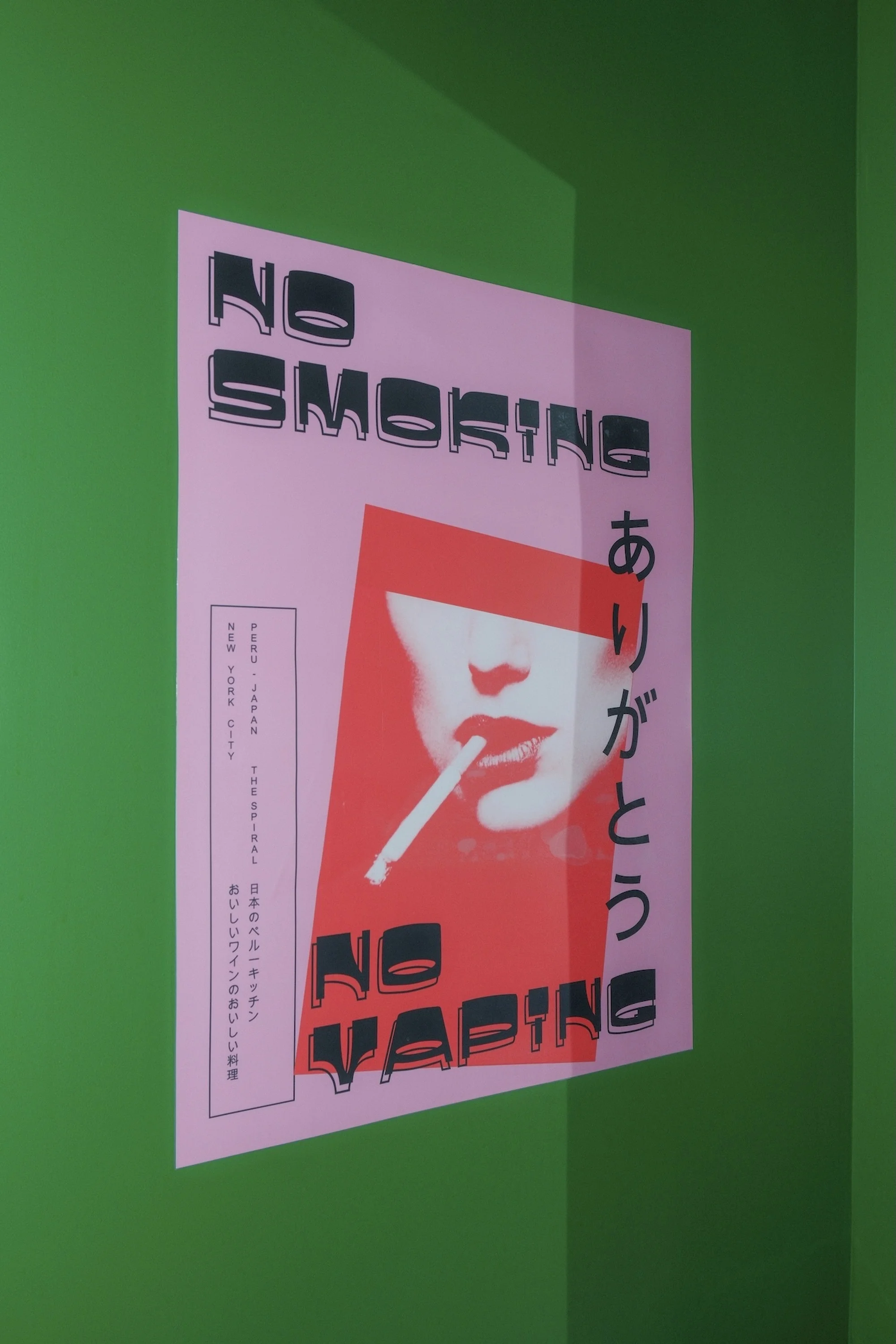 A pink poster on a green wall with 'No Smoking' and 'No Vaping' text, also featuring Japanese writing and a cigarette illustration.