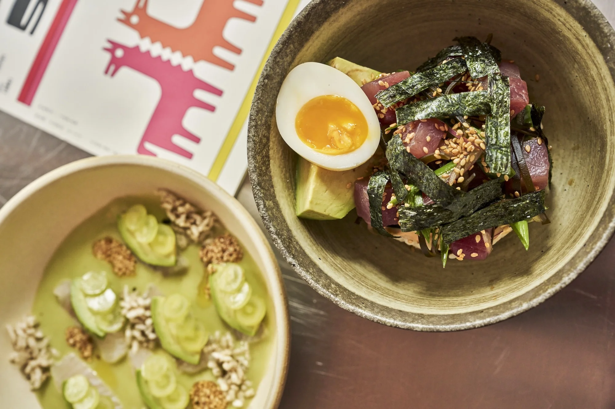 Overhead shot of two bowls filled with vibrant culinary creations: one with sliced avocado in green sauce, the other with tuna and seaweed.