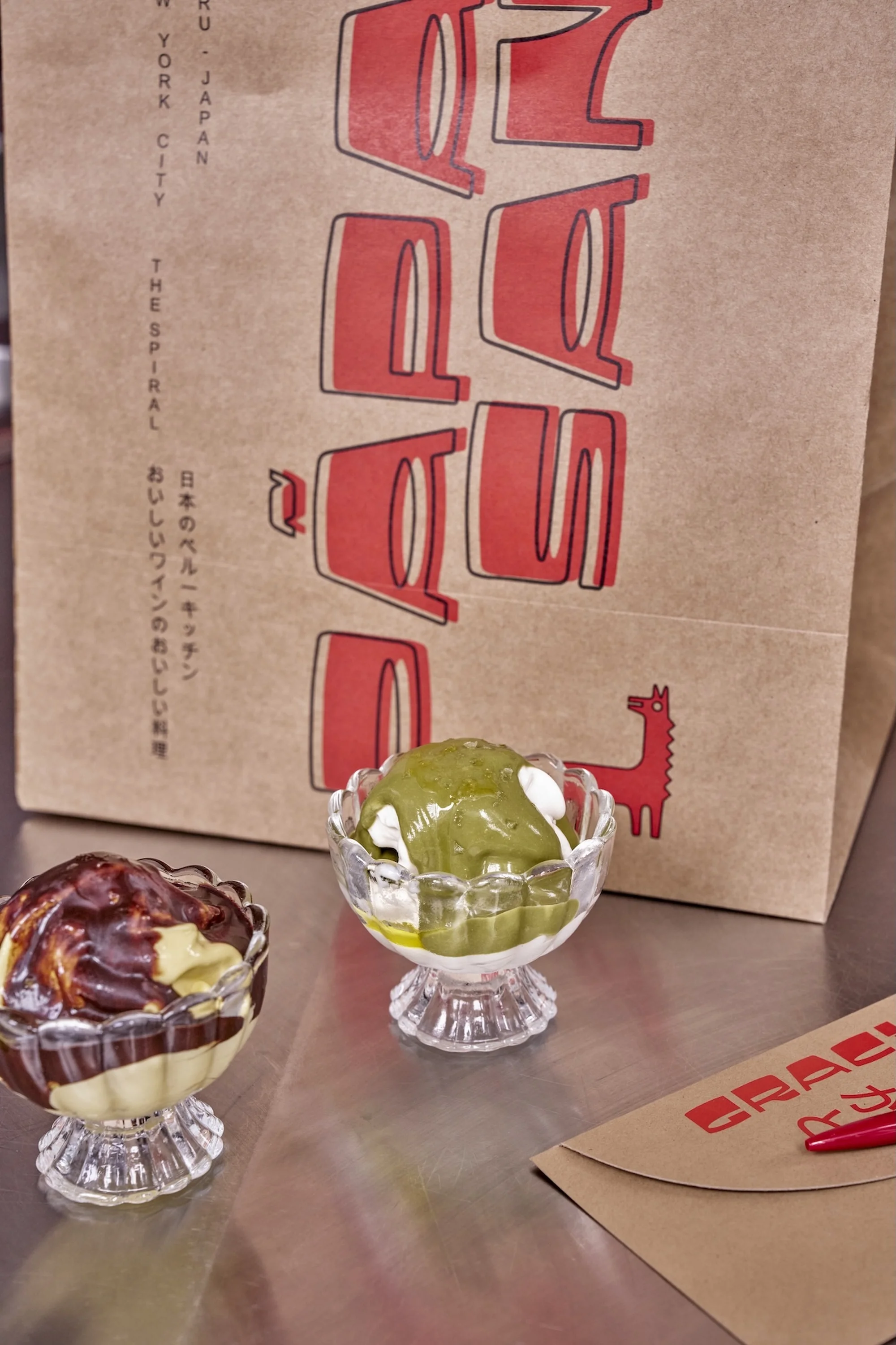 Two clear glass dessert cups filled with ice cream and toppings sit on a metal surface with a branded paper bag behind them.