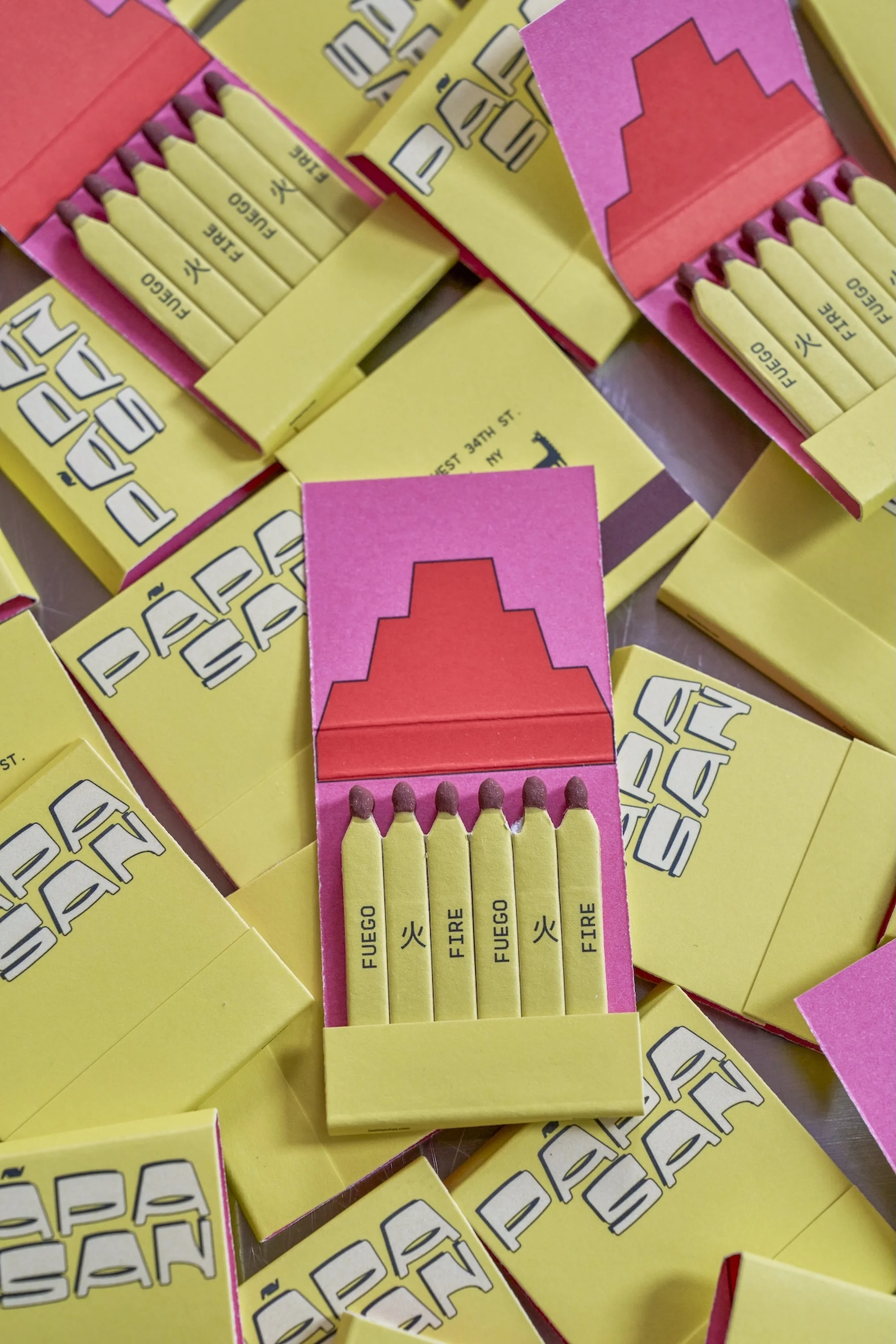 Arrangement of matchbooks featuring 'Fuego' and 'Fire' texts, set against a neutral metallic background, with dominant colors of yellow and pink.