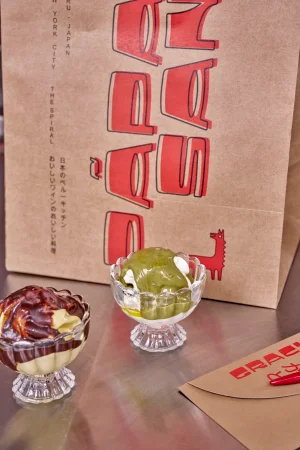 Two glass bowls of soft serve ice cream with sauce in front of a branded brown paper bag for Papa San
