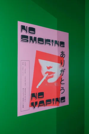 A pink graphic poster with black text reading NO SMOKING and NO VAPING mounted on a vibrant green wall corner.