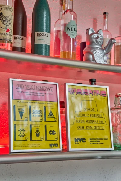 Shelves displaying liquor bottles, a silver lucky cat figurine, and two framed informational signs with yellow and pink designs.