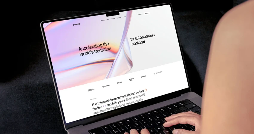 Person types on a laptop displaying the Coders website, which promotes the transition to autonomous coding. The laptop sits on a black surface.