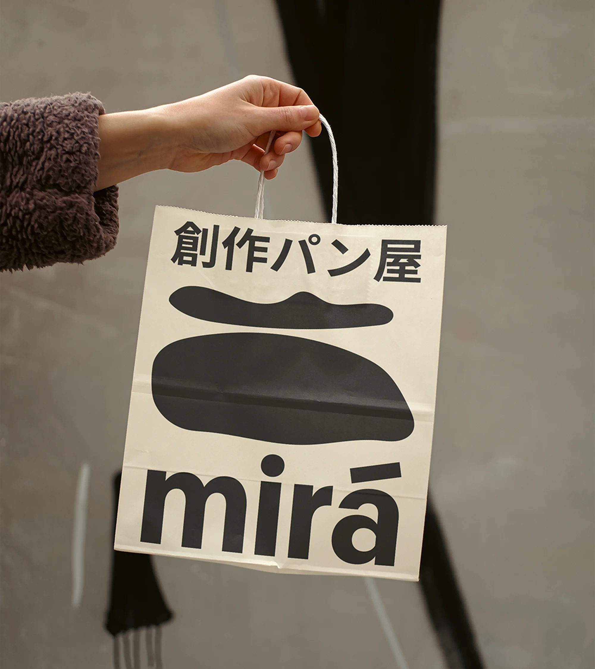 Close-up of a hand holding a paper bag with Japanese writing and black graphic shapes against a blurred neutral background.