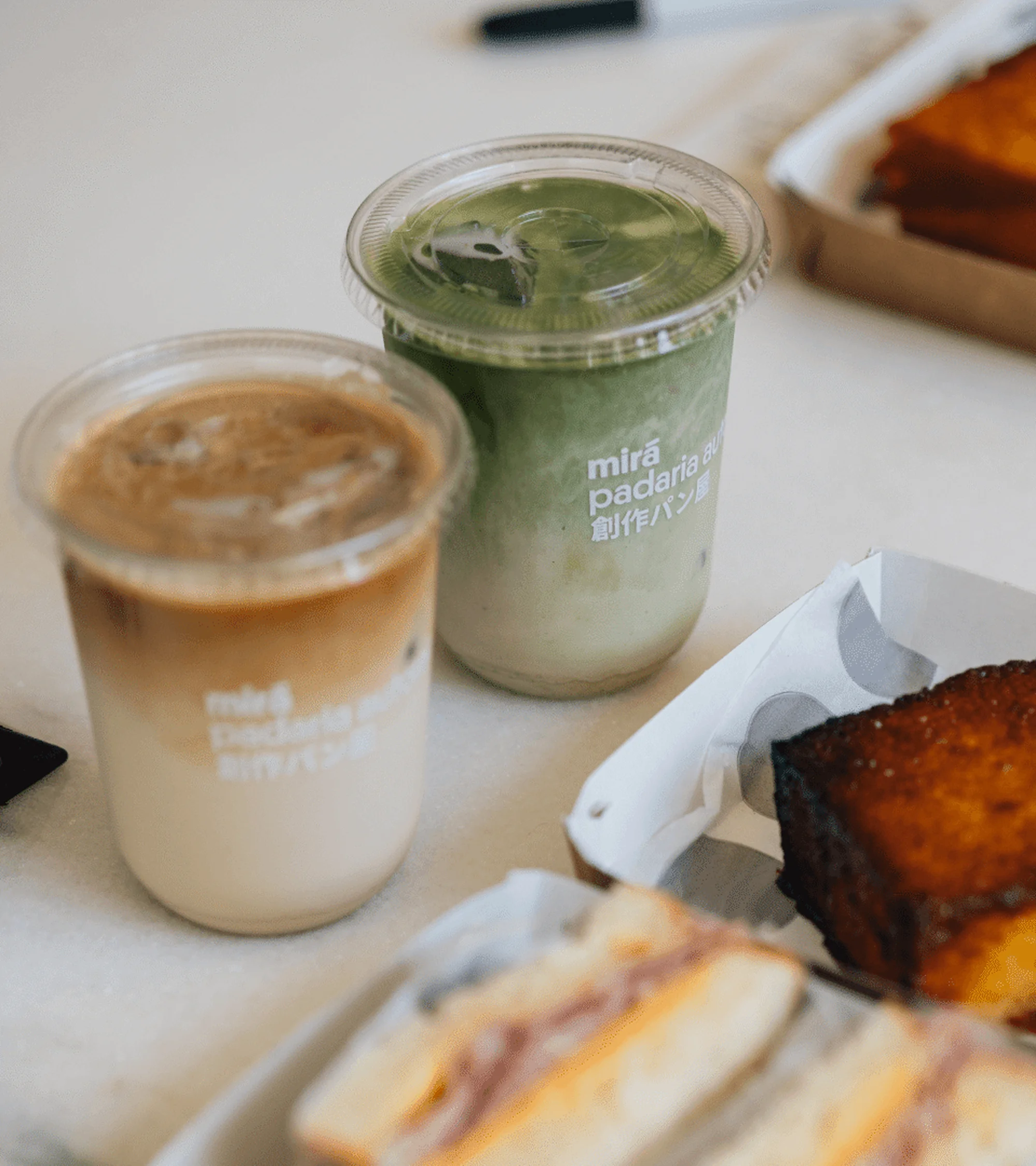 Two iced drinks, matcha and coffee, stand near sandwiches on a white surface, highlighted by soft lighting and focus on culinary presentation.