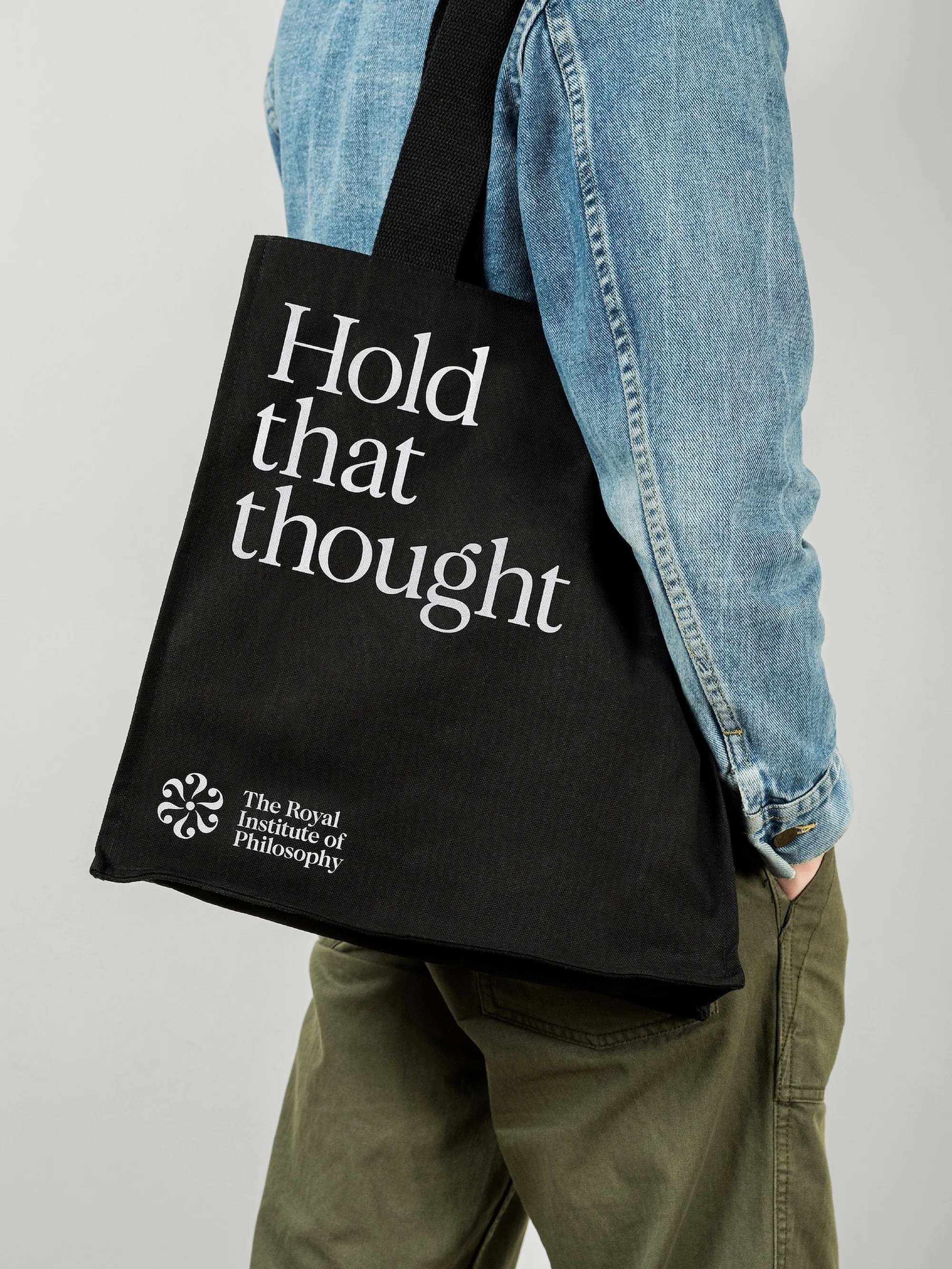 Person carrying black tote bag with white text 'Hold that thought,' wearing denim jacket and olive green pants on neutral backdrop.