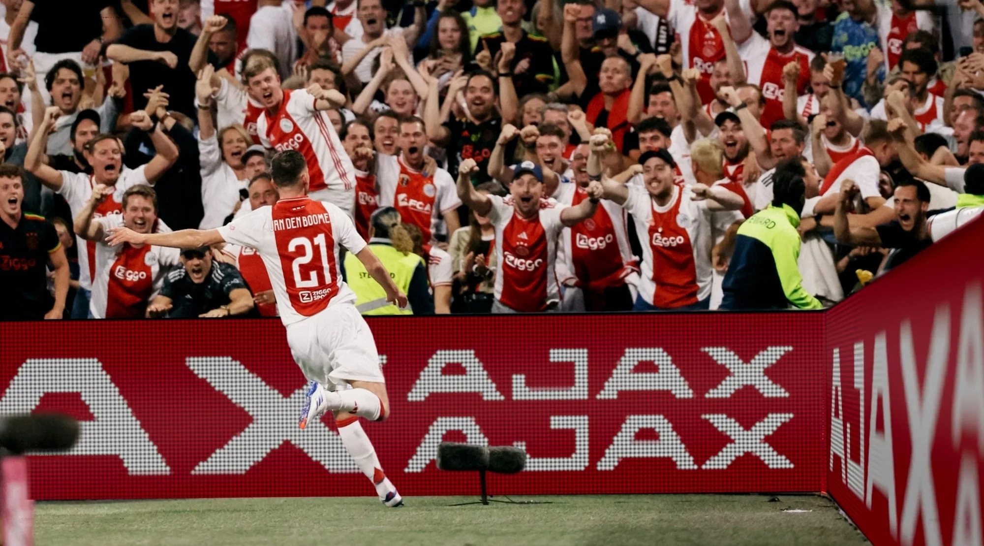 Soccer player wearing number 21 jumps with arms outstretched in front of cheering fans in red and white jerseys at a stadium.