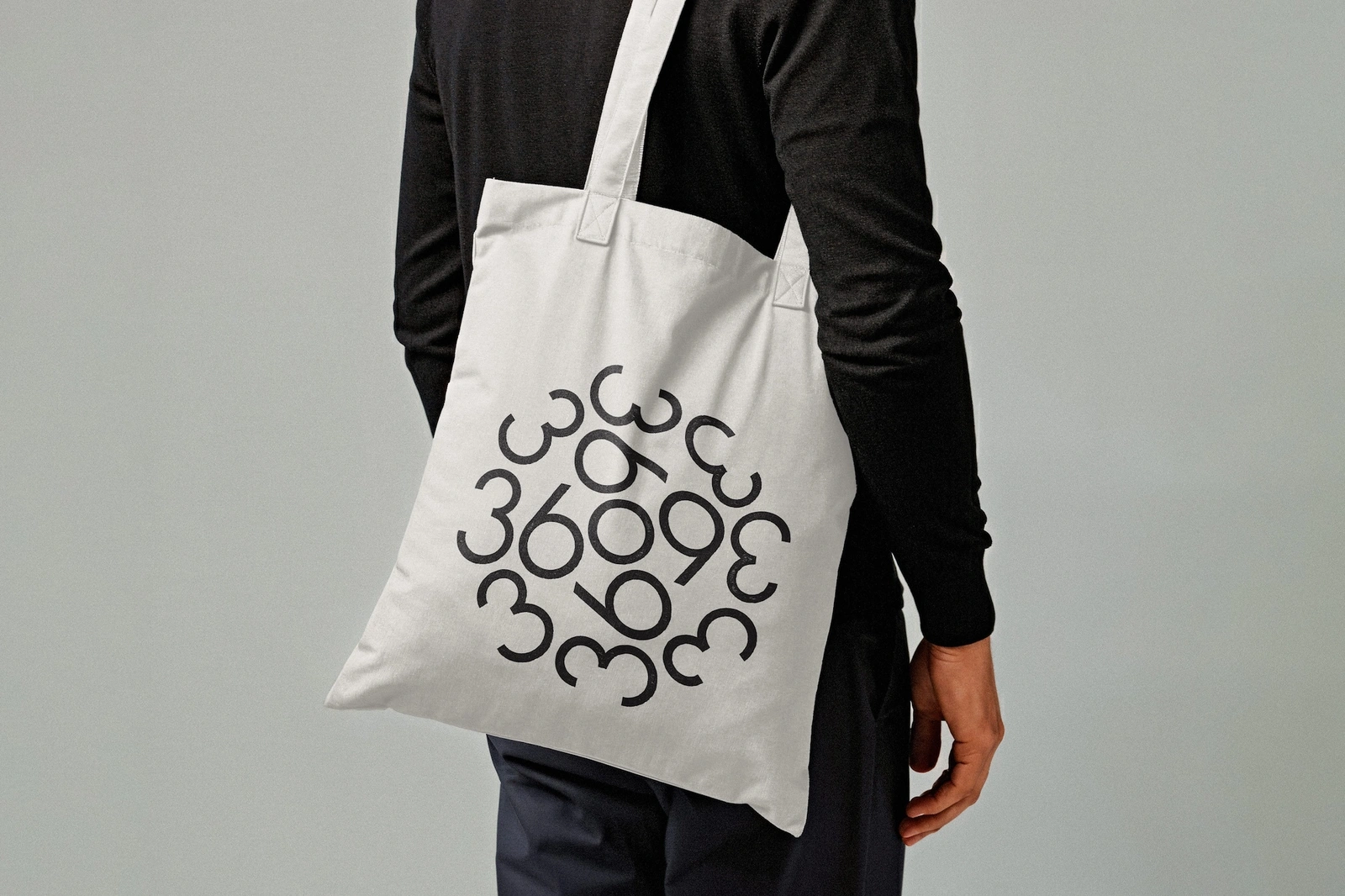Person wearing a black sweater and dark trousers carrying a white canvas tote bag with black typographic logo design.
