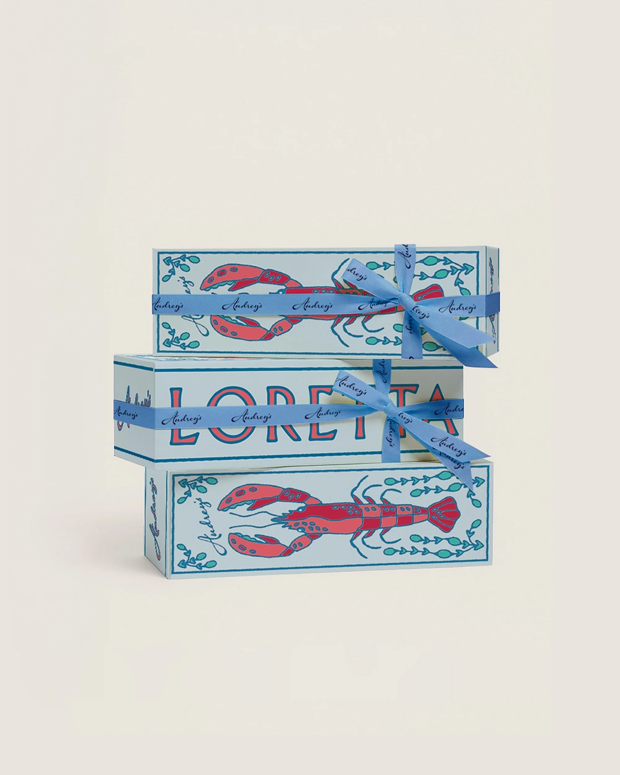 A stack of three rectangular gift boxes with an illustration of a lobster and the word 'Loretta', tied with a blue ribbon.