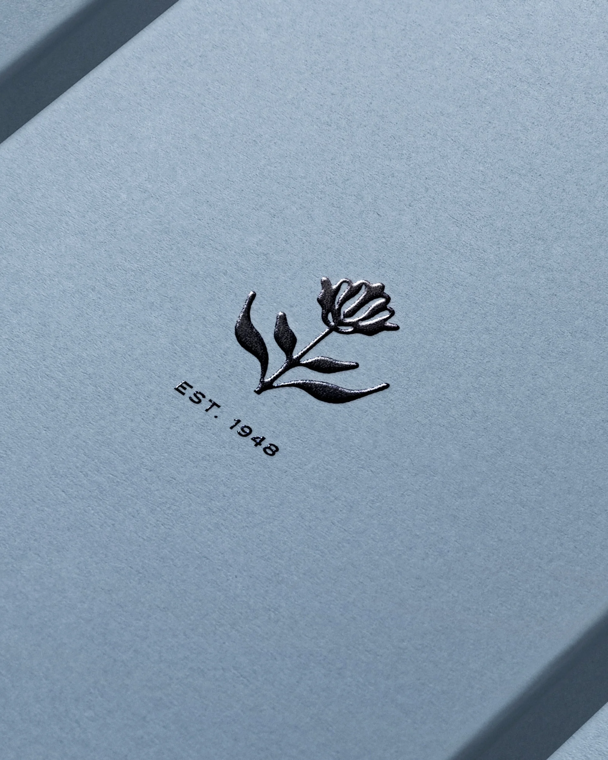 Close-up of a light blue card with a dark grey floral emblem and the text 'EST. 1948' printed below, conveying a sense of classic branding.