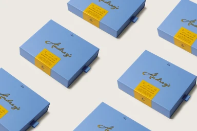 An overhead diagonal pattern of light blue chocolate gift boxes with yellow belly bands on an off-white background.
